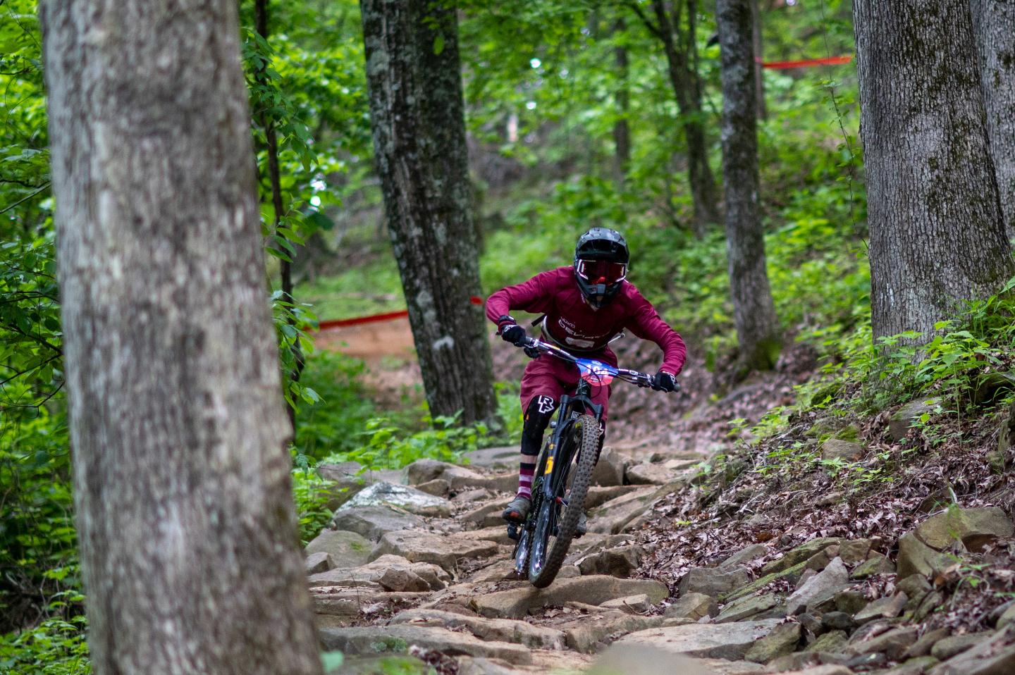 A cyclist navigates a rugged forest trail, surrounded by green trees.