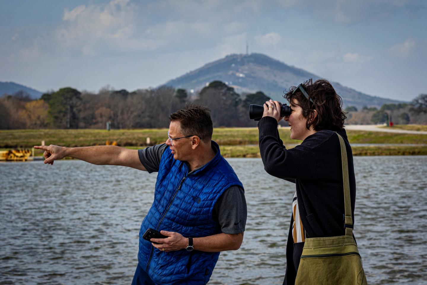Two people by water, one pointing and the other using binoculars. Mountain in the background.
