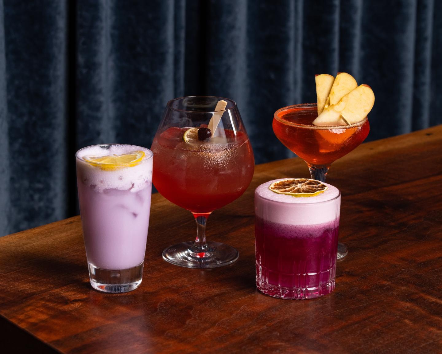 Four colorful cocktails on a wooden table with a dark curtain backdrop.