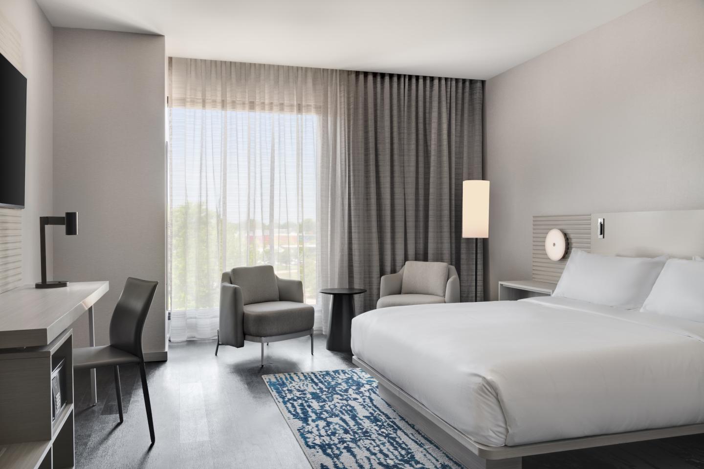 Modern hotel room with king bed, desk, chairs, and large window with curtains.