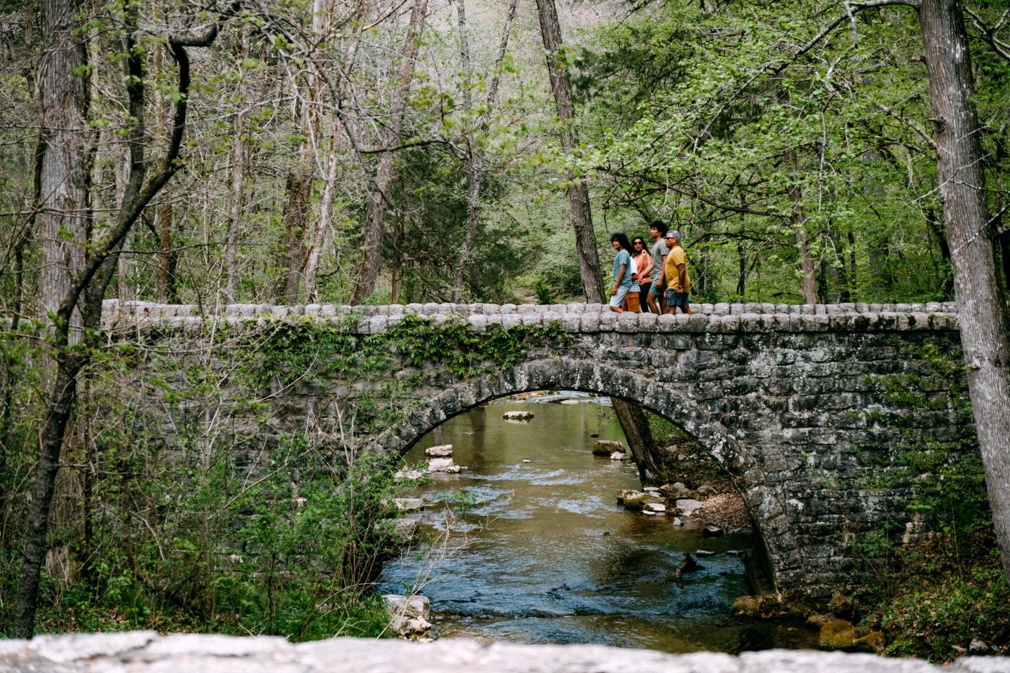 Cyclist in orange crosses a stone bridge over a wooded creek.