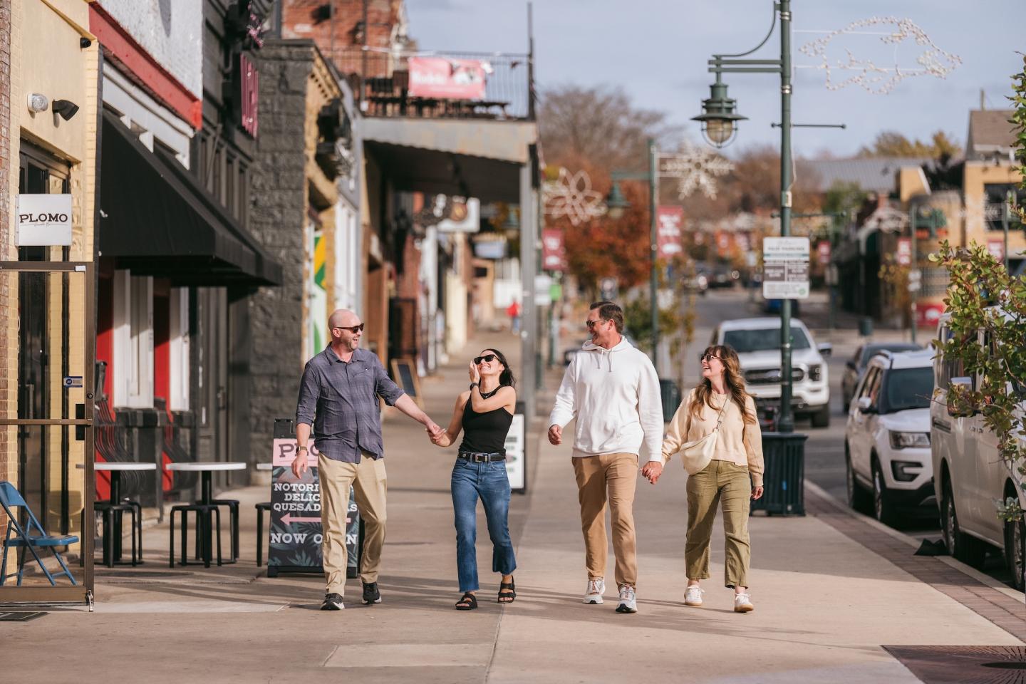 Four people walking on a sunny street lined with shops and cars.