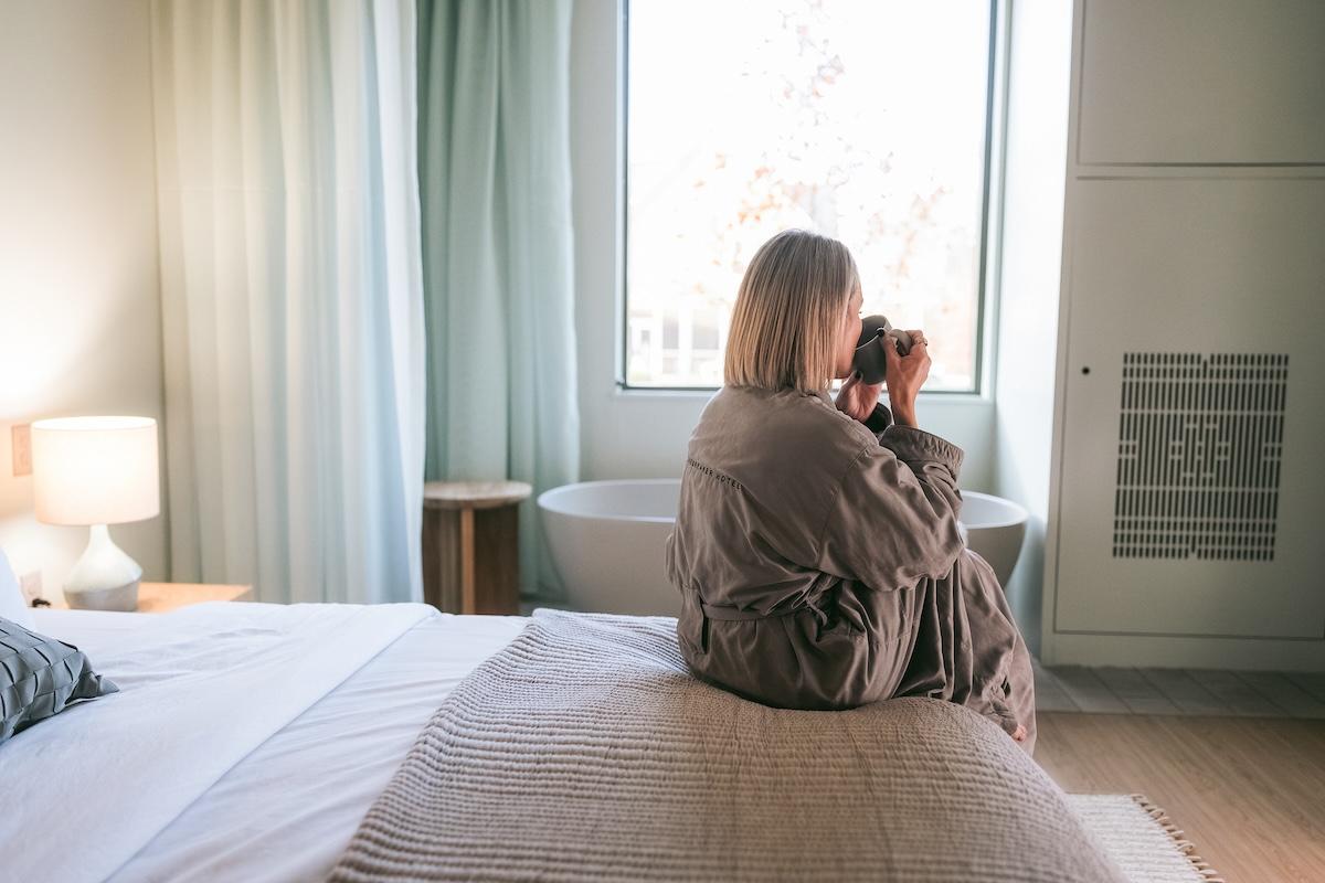 Woman sitting on a bed, drinking coffee by a window.