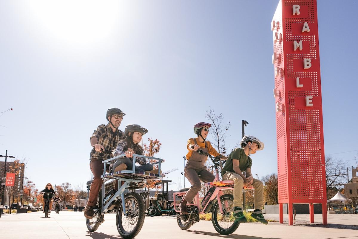 Cyclists ride in a sunny urban area, passing a tall red sign.