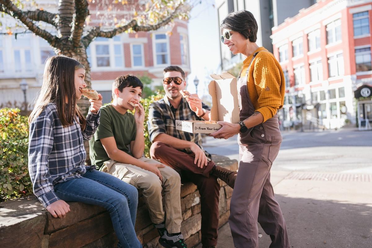 Street scene with a woman handing food to three seated teens.