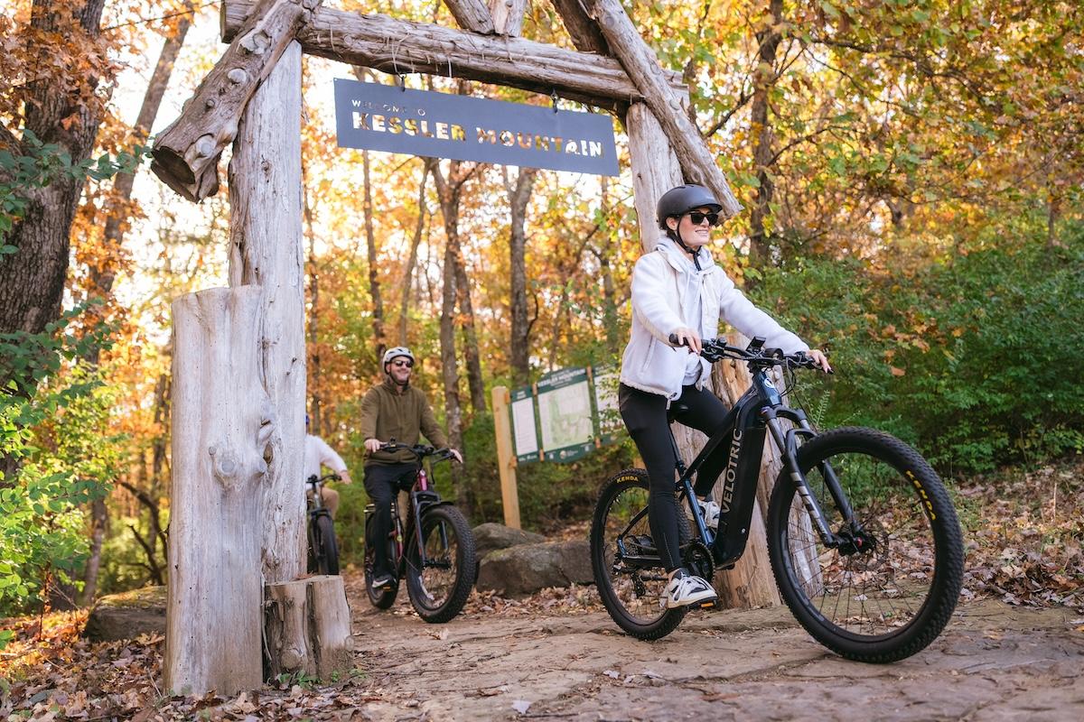 Cyclists ride under a wooden arch in an autumn forest setting.