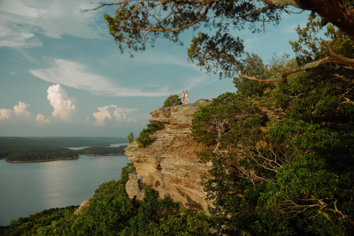 Cliffside view with two people overlooking a lake under a cloudy sky.