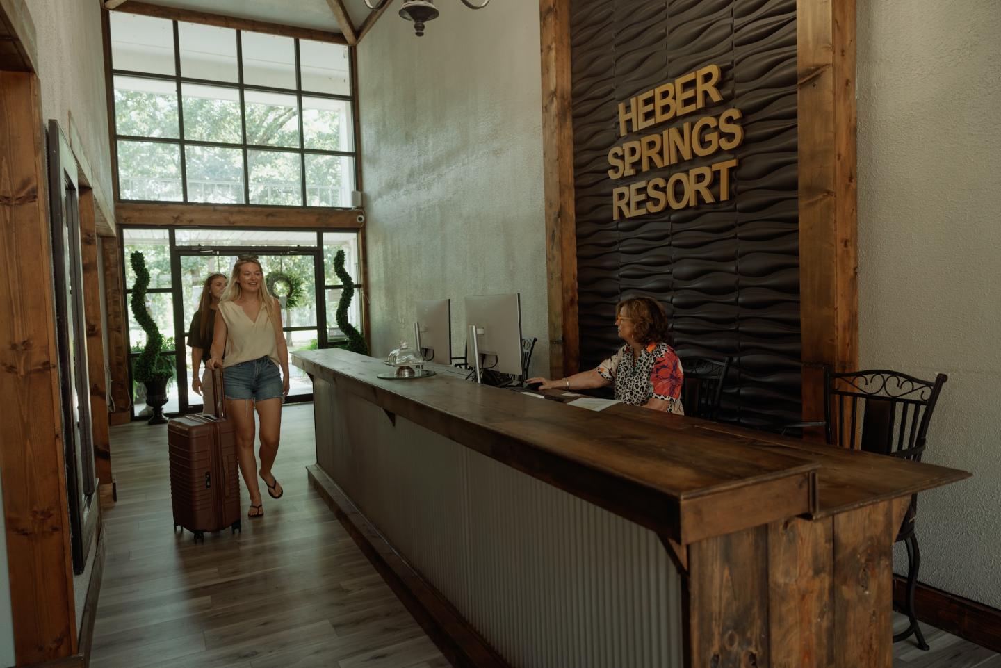 Woman with suitcase at hotel reception desk, wood decor, large windows.