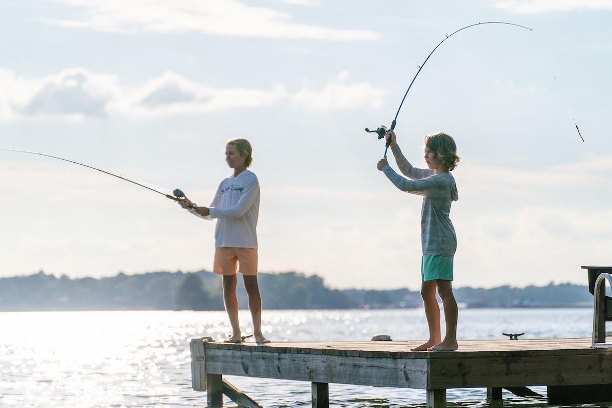 Two children fishing on a sunny dock by a lake.