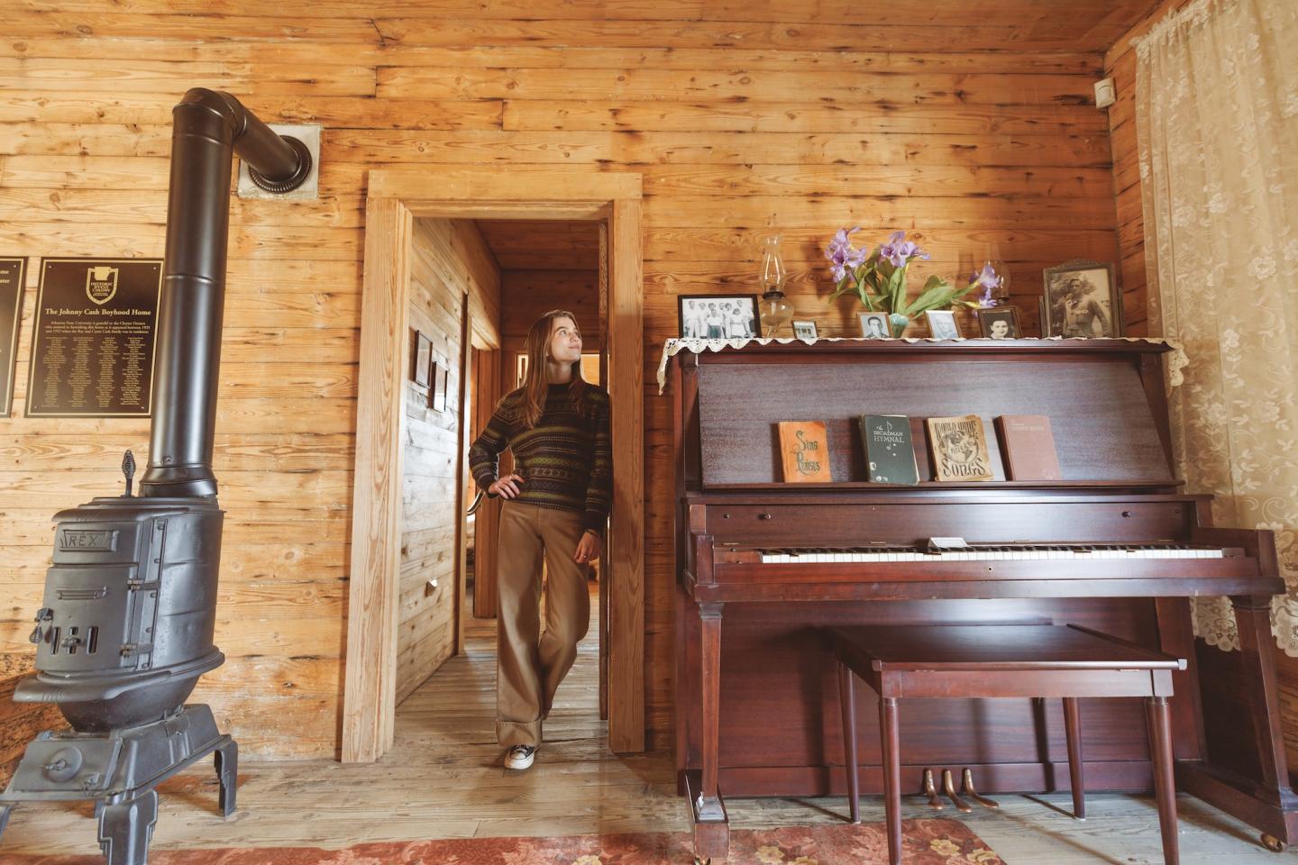 Cozy wooden room with a piano, wood stove, and a person by the door.