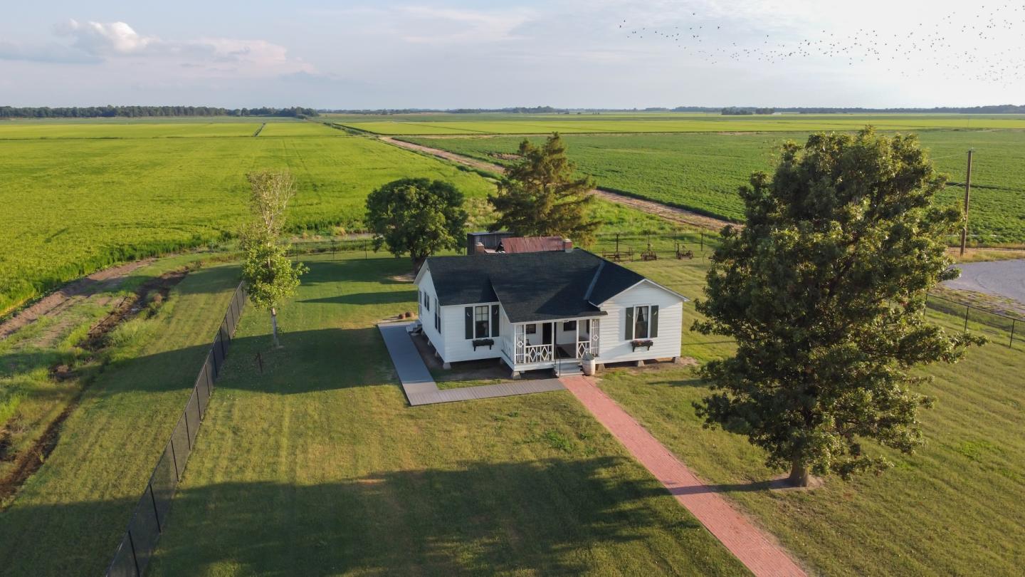 Small white house with black roof in a vast green field, surrounded by trees.