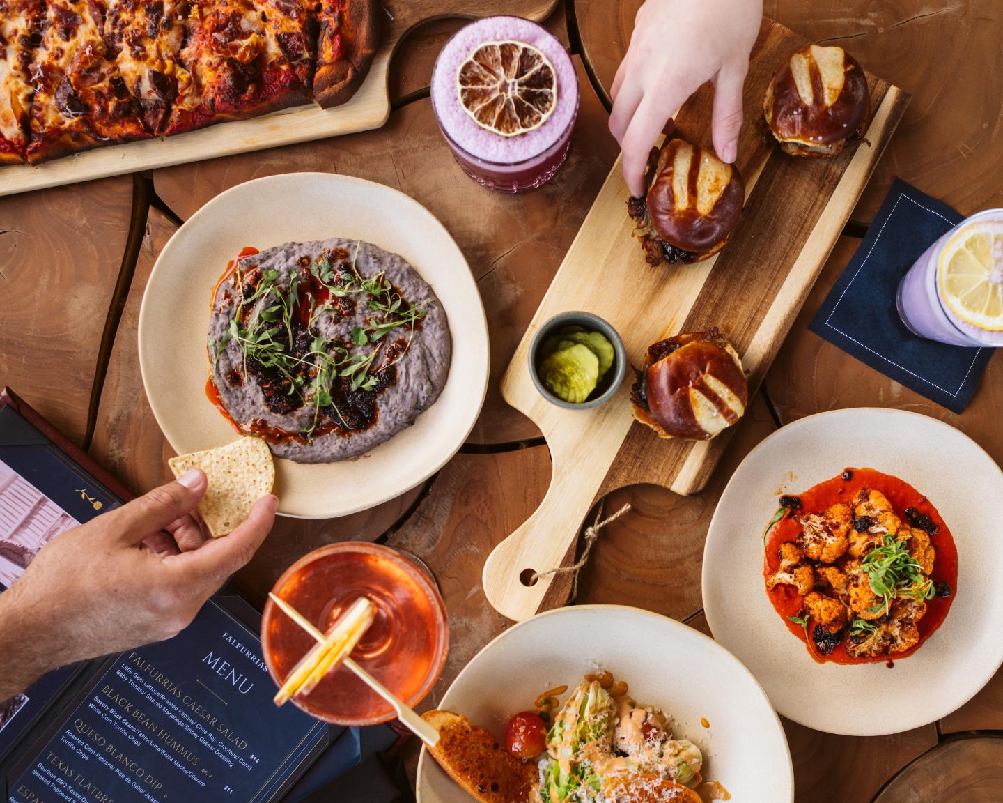 A table with assorted dishes, hands holding bread and drinks in various colors.