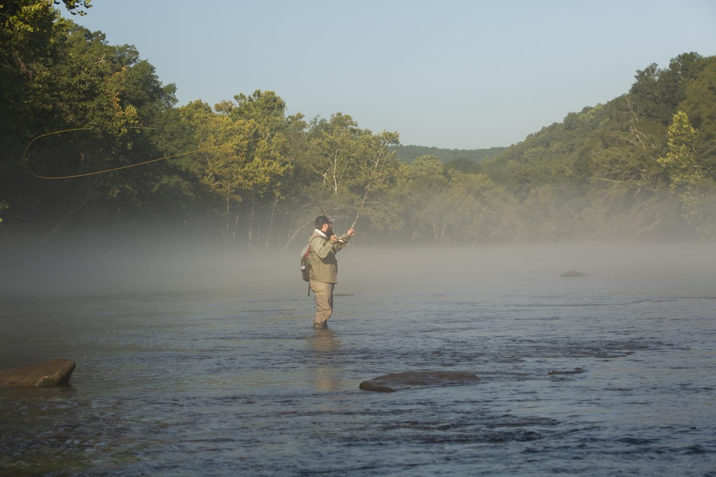 Fly fisher wading in misty river, surrounded by trees and hills.