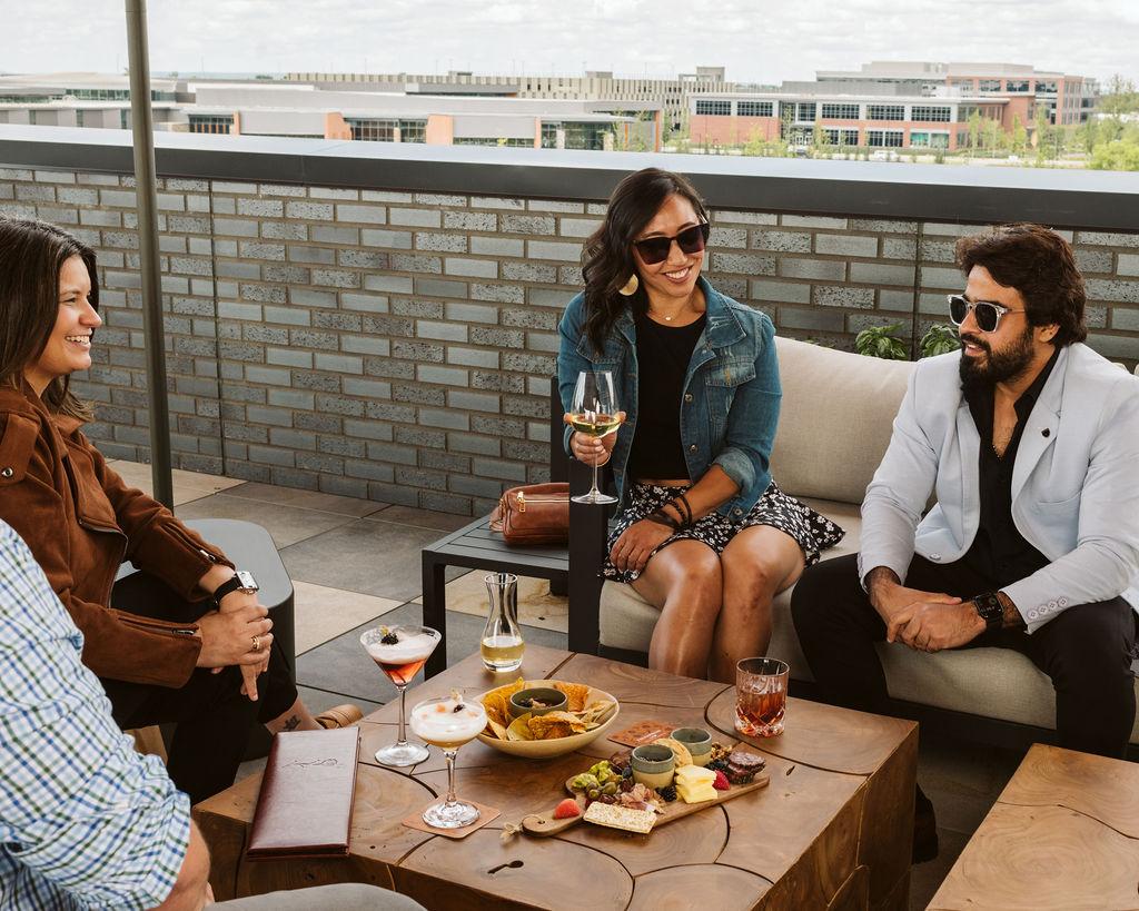 Four people sitting on a rooftop patio, chatting and enjoying drinks and snacks.