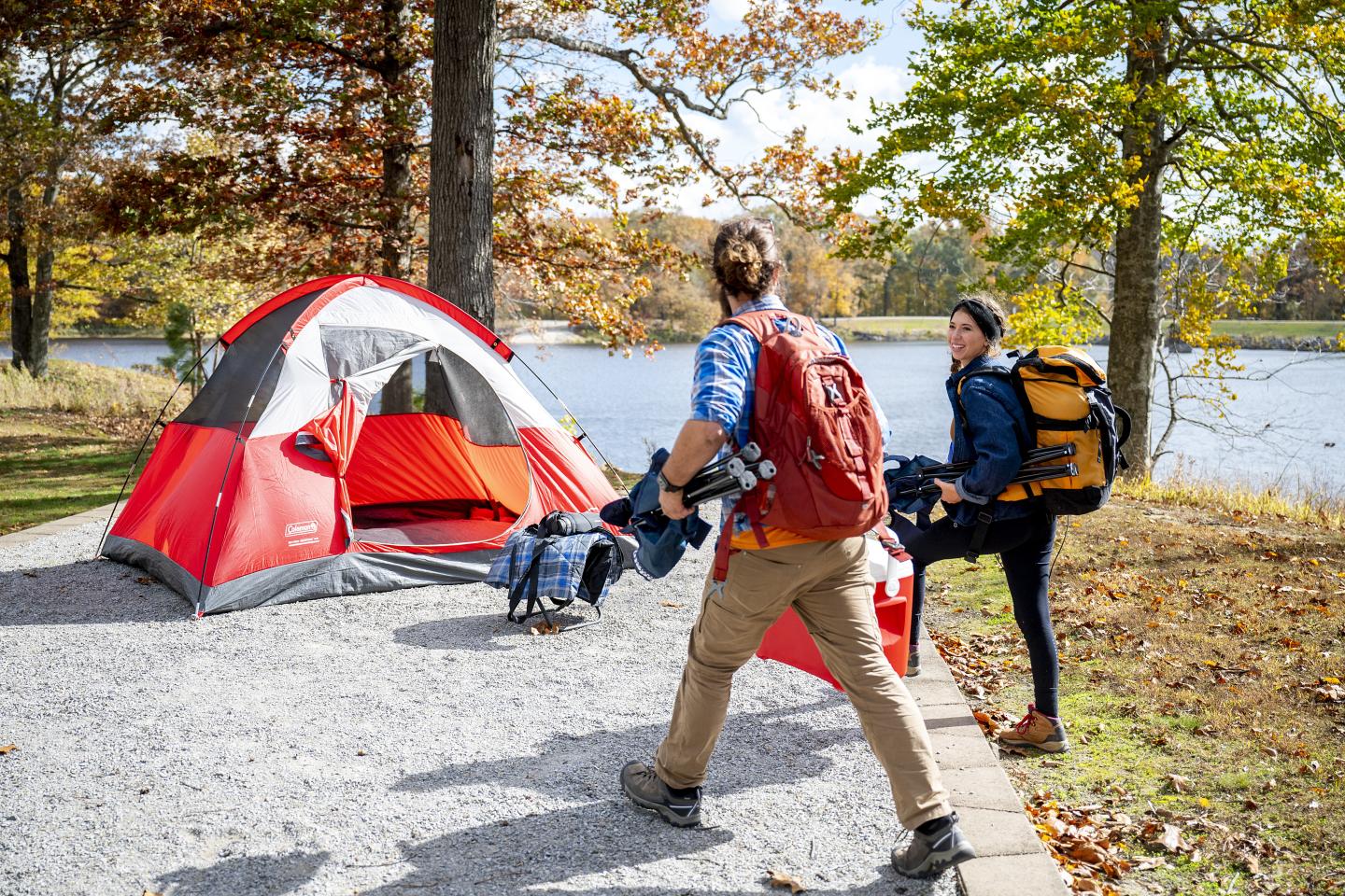 Two hikers walk past a red tent beside a lake in autumn.