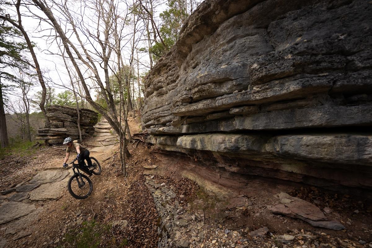 Mountain biker riding beside a layered rock cliff on a forest trail.