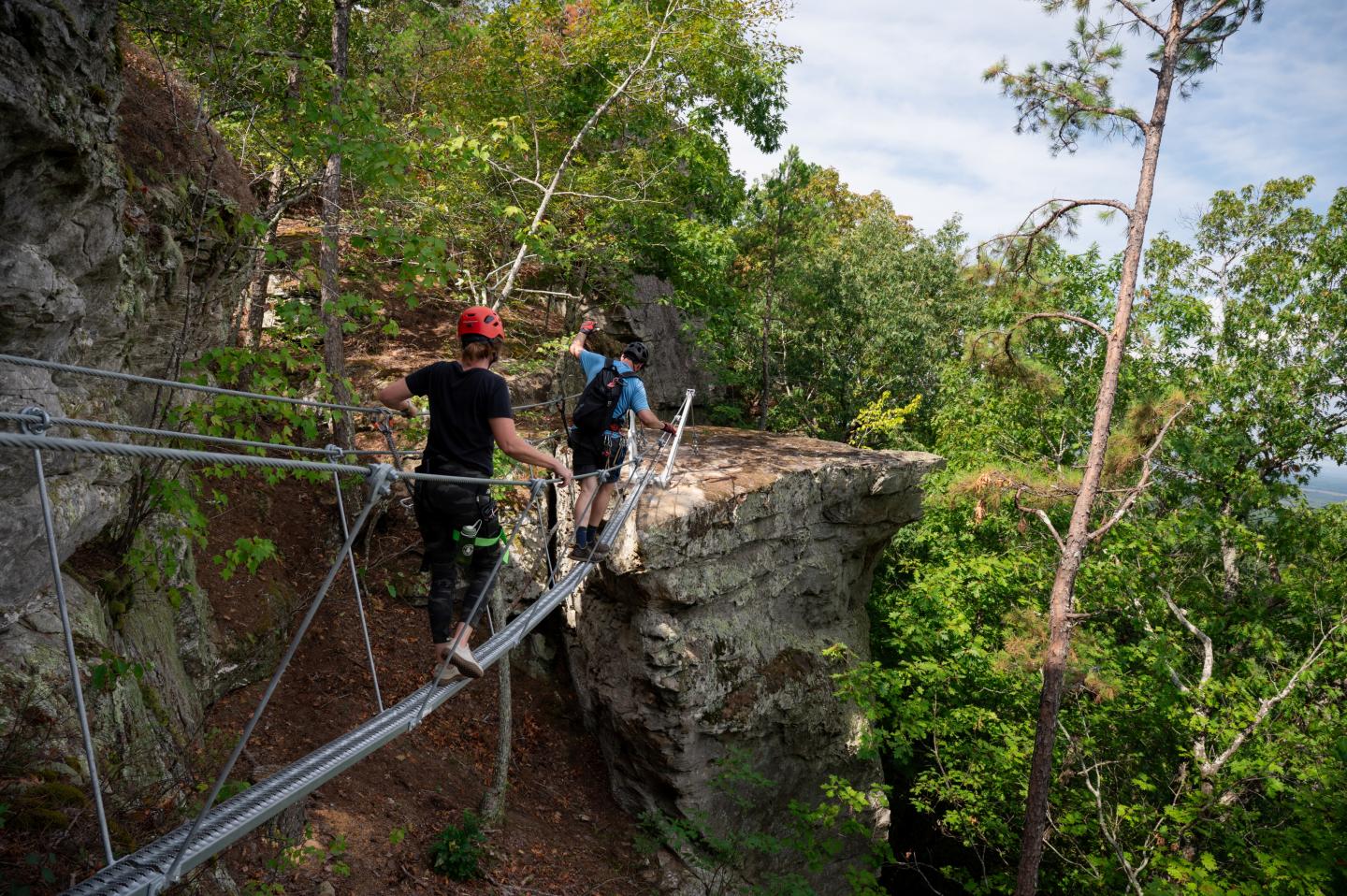 Hikers crossing a narrow suspension bridge along a forested cliff trail.