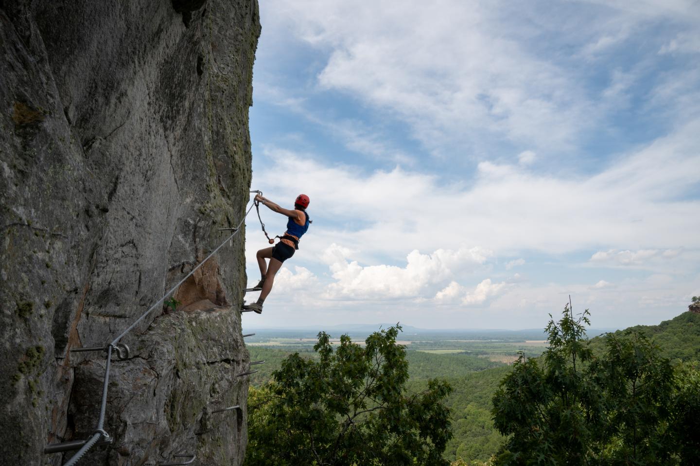 Rock climber ascending a steep cliff above a green valley under a cloudy sky.