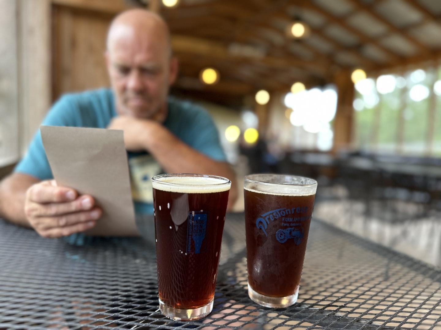 Two pints of dark beer on a patio table, with a man using a tablet behind them.