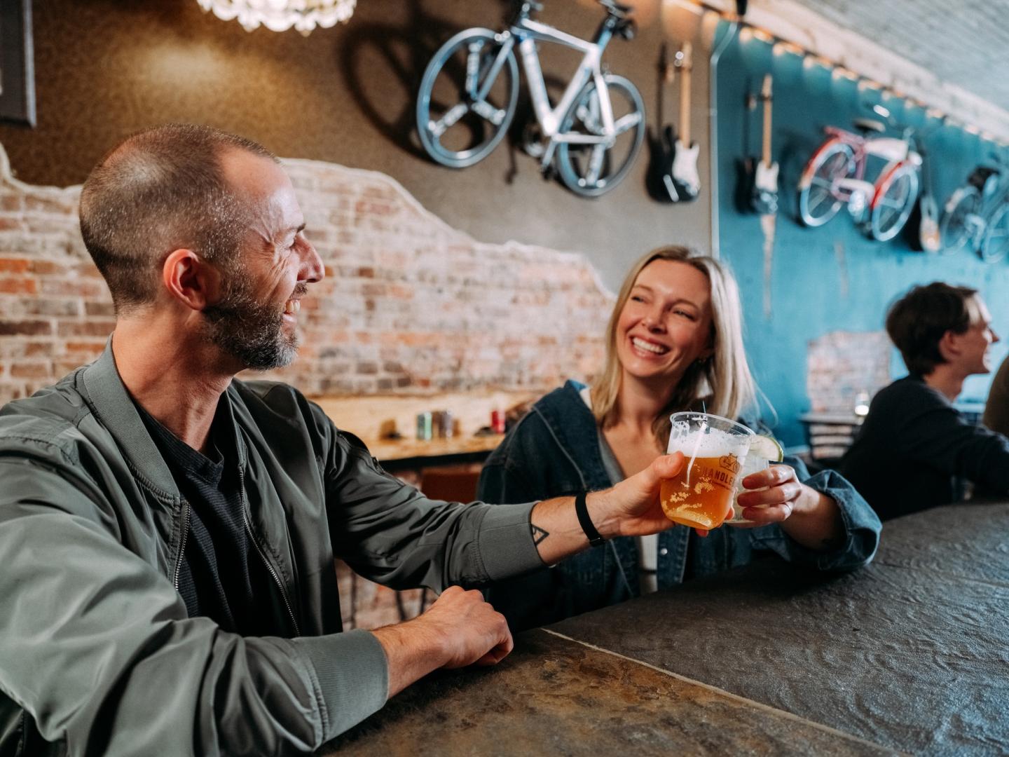 Two friends clink beers at a rustic brewery bar with bicycles overhead.