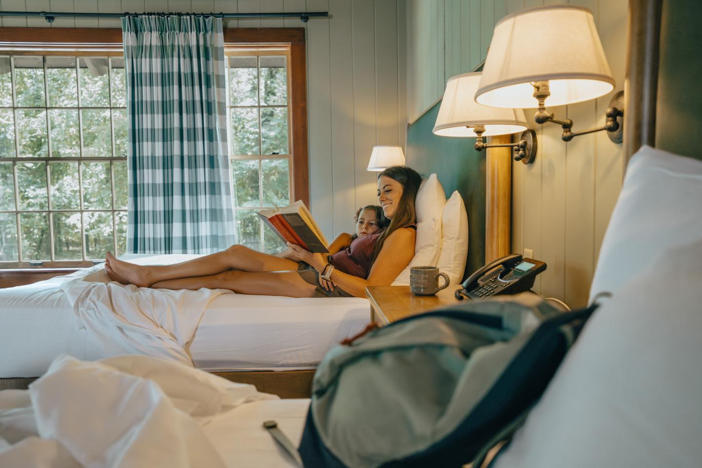 Person relaxing on a hotel bed, reading a book by a window.