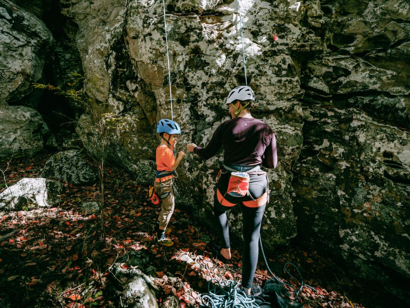 Hiker in helmet and backpack standing beside a rocky cliff in a forest.