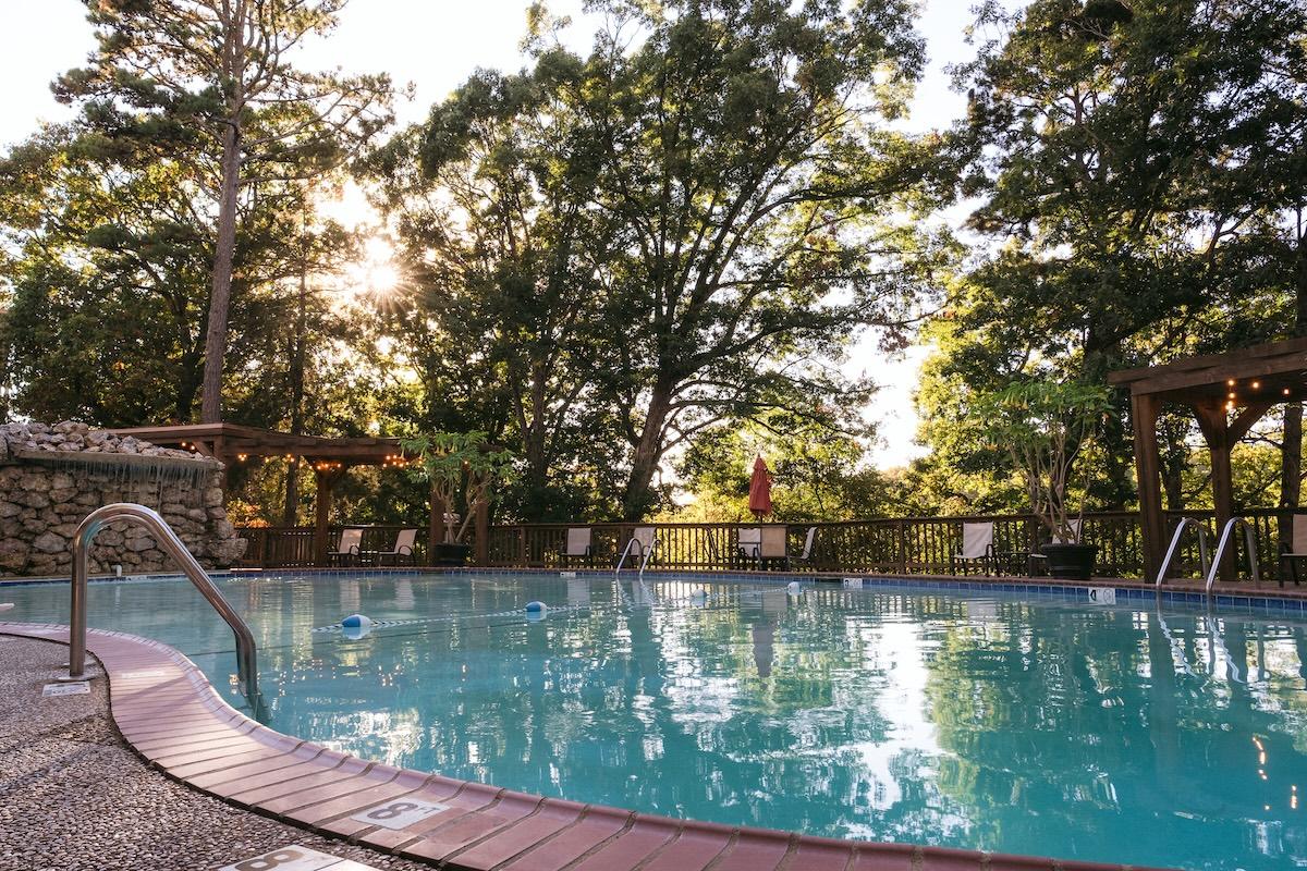 Outdoor swimming pool surrounded by trees, with sunlight filtering through.