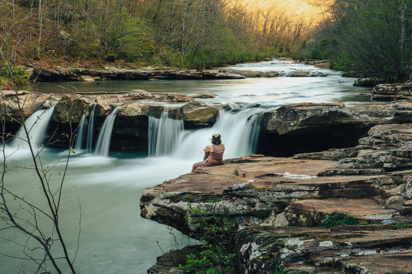 Person sitting on rocks beside a small waterfall in a forest stream.
