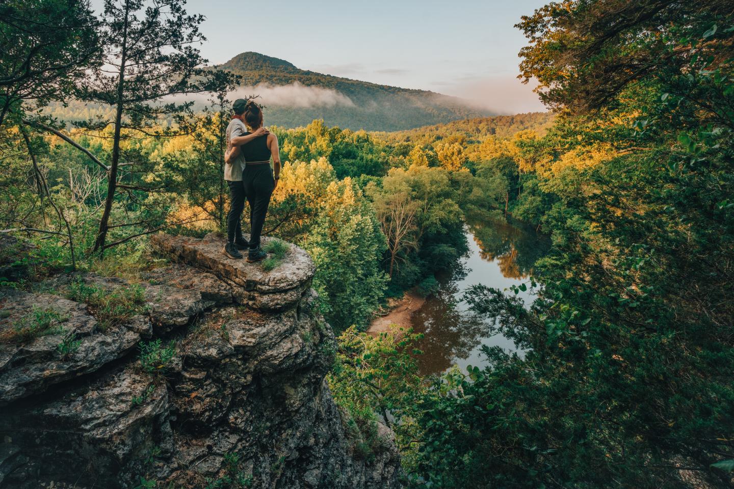Hiker standing on a rocky cliff above a forested river valley at sunset.