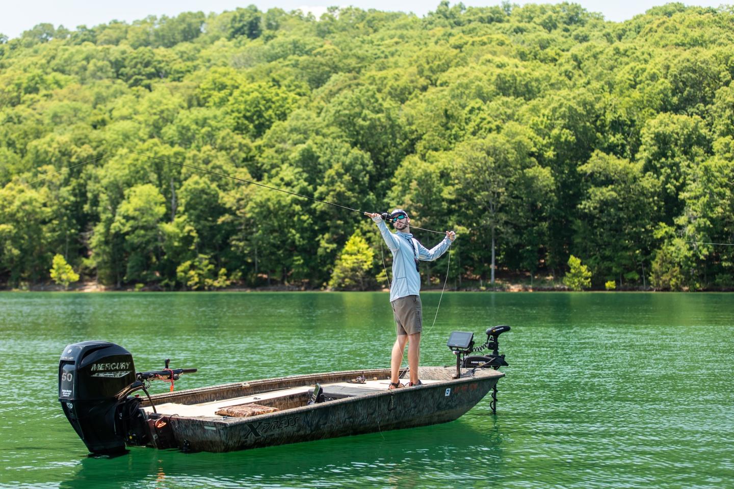 Man fishing from a small boat on a green lake, surrounded by forest.