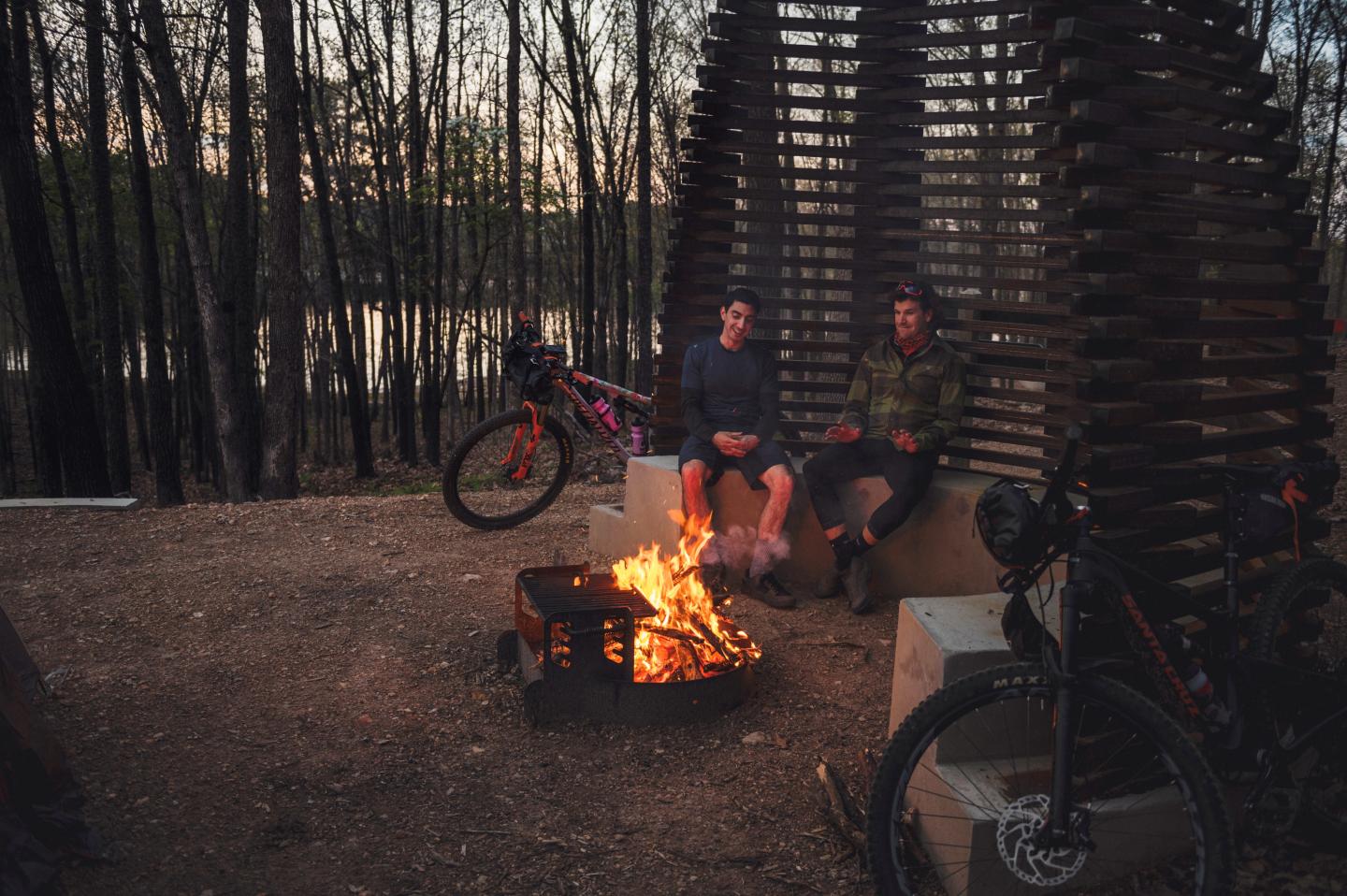 Two cyclists warm by a campfire beside a log cabin in a forest.