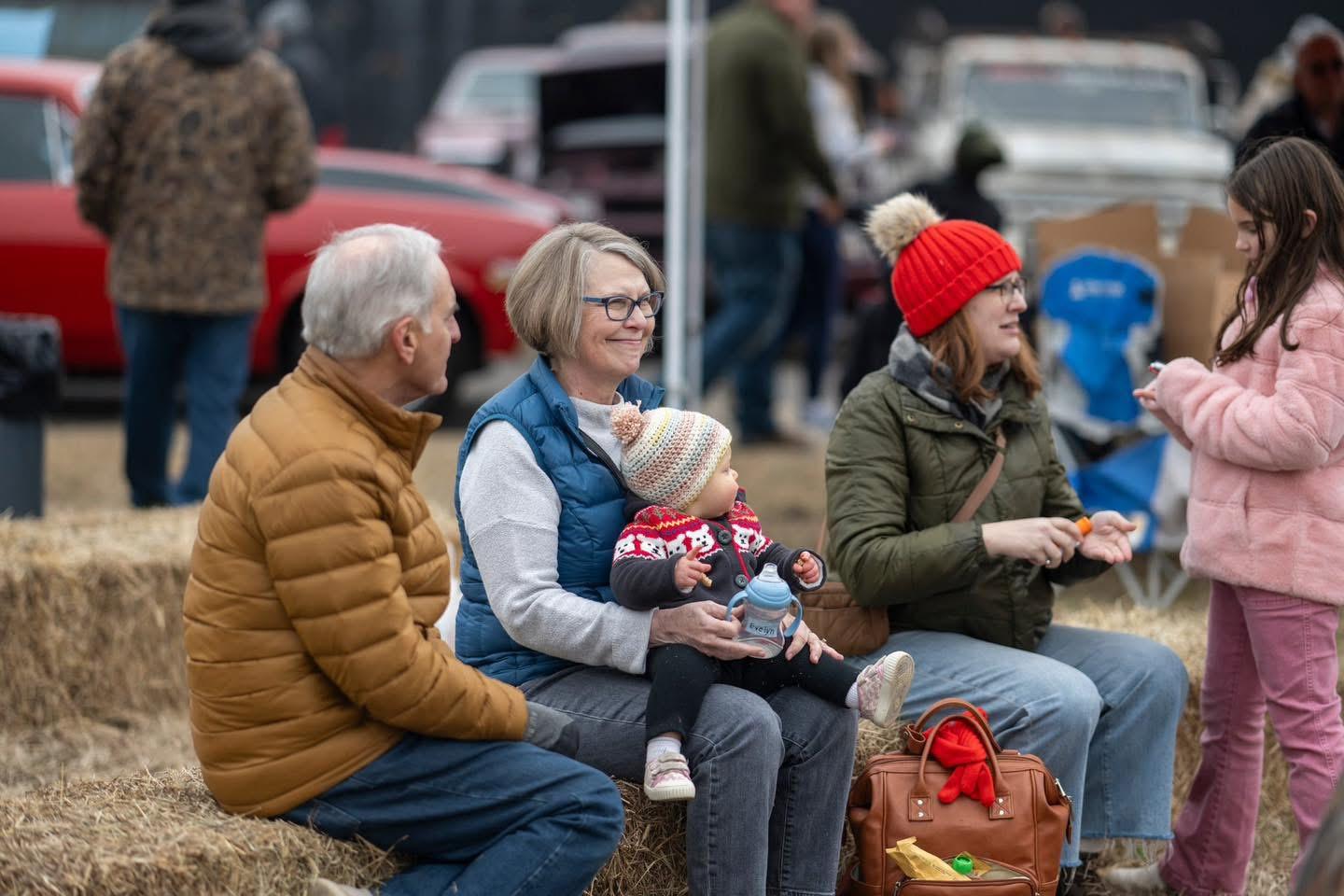 Spectators in winter clothing sit on hay bales at an outdoor event.