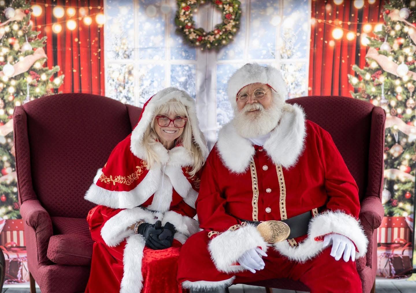 Santa and Mrs. Claus sit in red chairs before festive Christmas decorations.