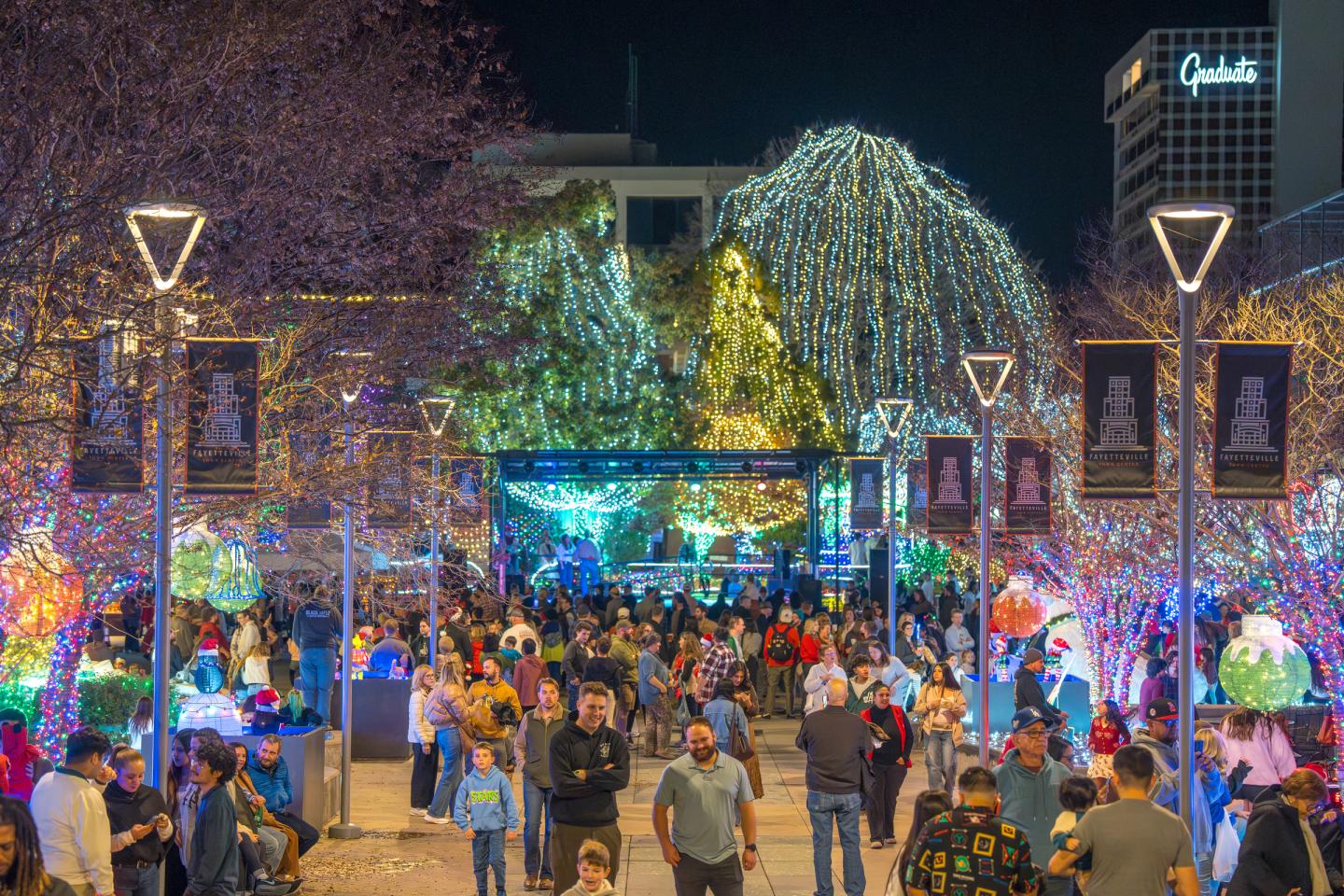 Crowded nighttime festival street with colorful lights and illuminated trees.