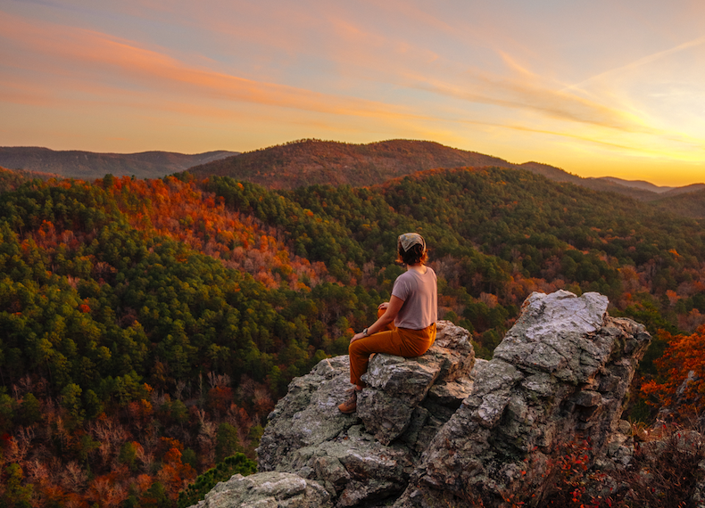 Hiker seated on rocky overlook, watching a sunset over autumn-covered hills