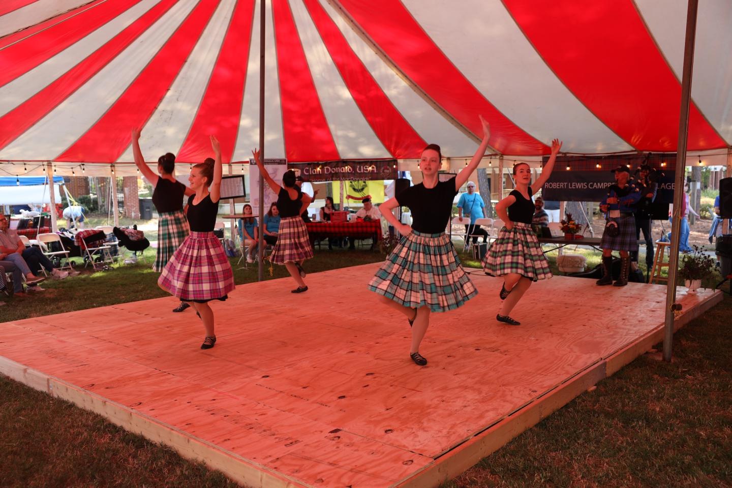 Dancers in tartan skirts perform on a stage beneath a red-and-white striped tent.