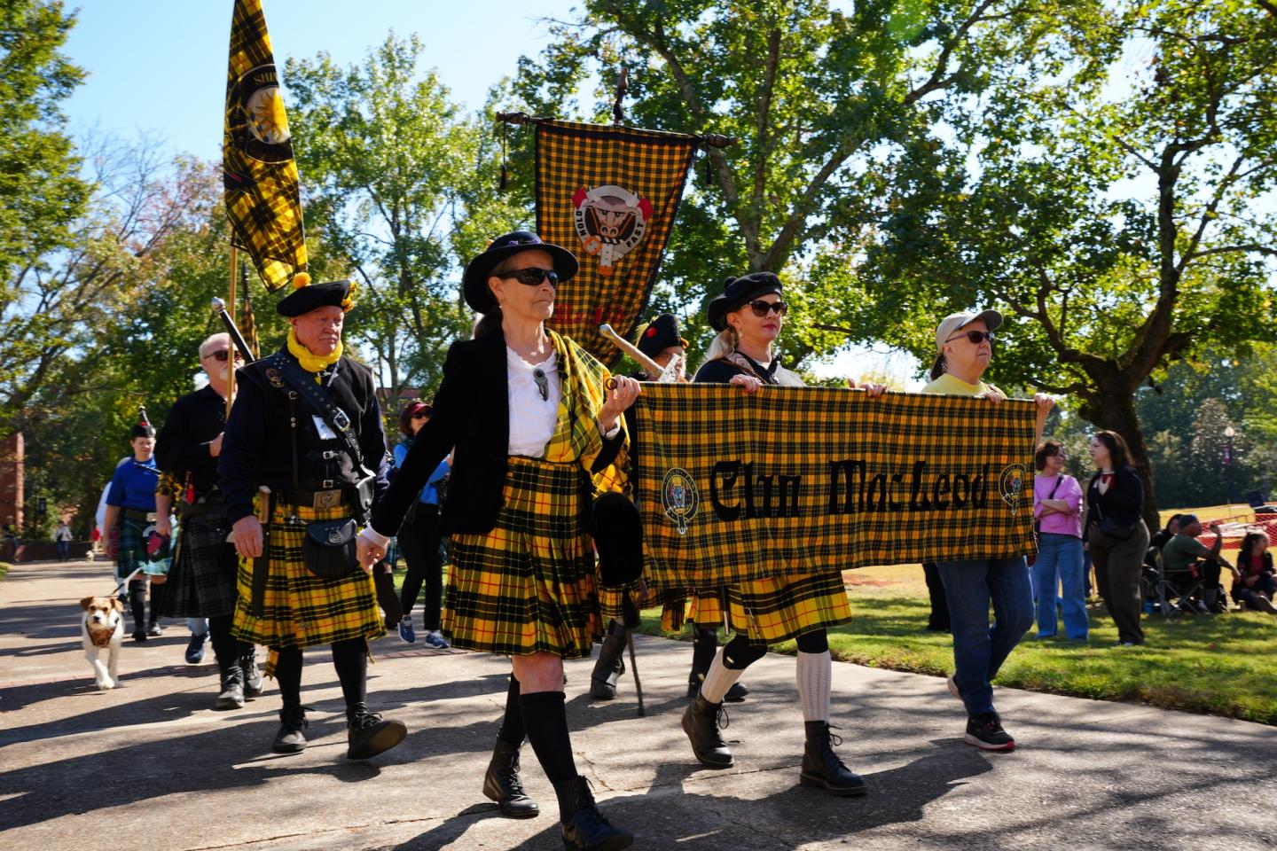 Bagpipe band members in yellow tartan kilts marching in a sunny park.