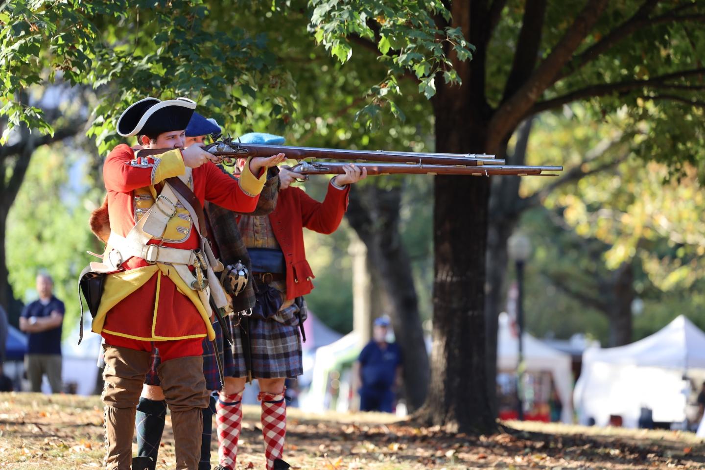 Two children in colorful costumes aim a toy rifle in a park.