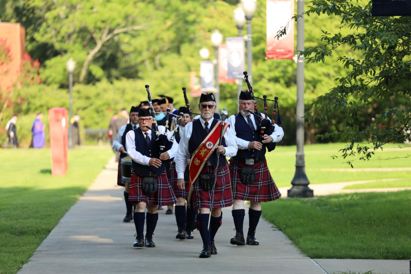 Four bagpipers in formal Scottish dress march along a park path.