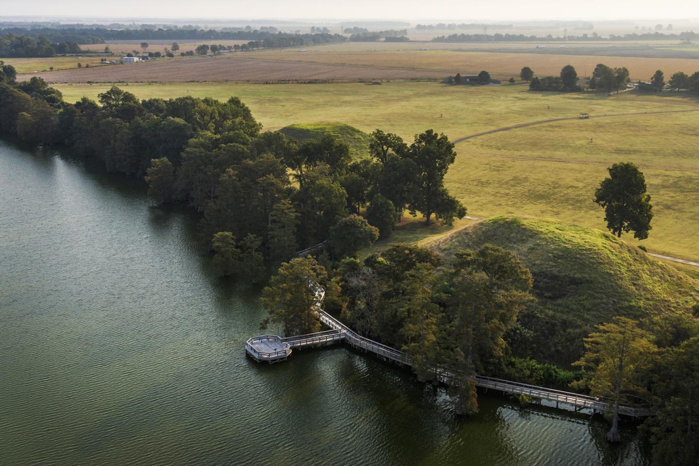 Aerial view of a tree-lined riverbank with a small dock and boat beside farmland
