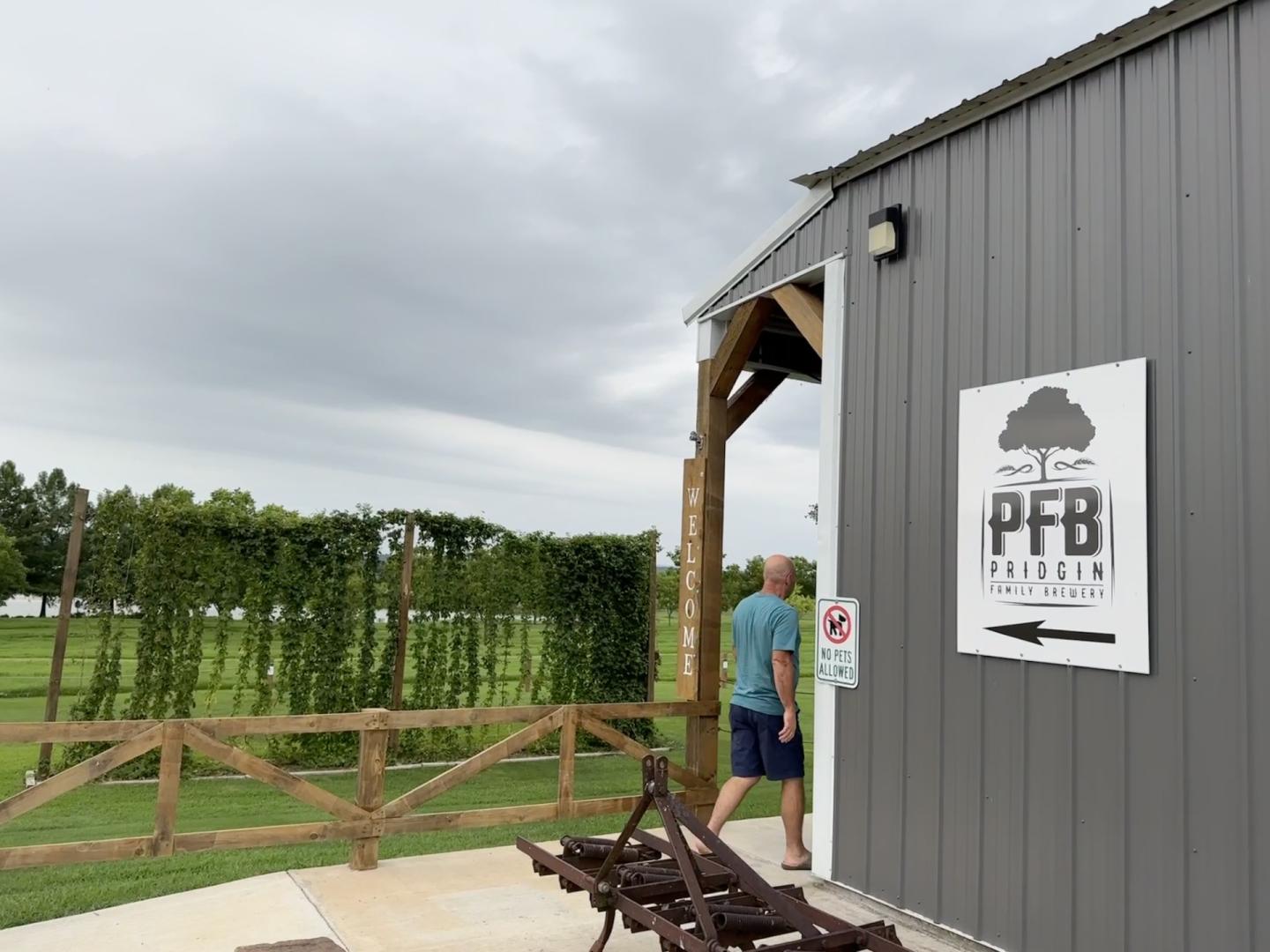 Gray barn with an HFB sign and arrow, beside a wooden gate and green field.
