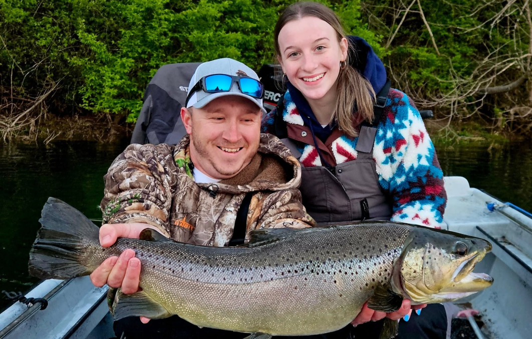 Smiling couple holding a large trout in a boat on a wooded river.