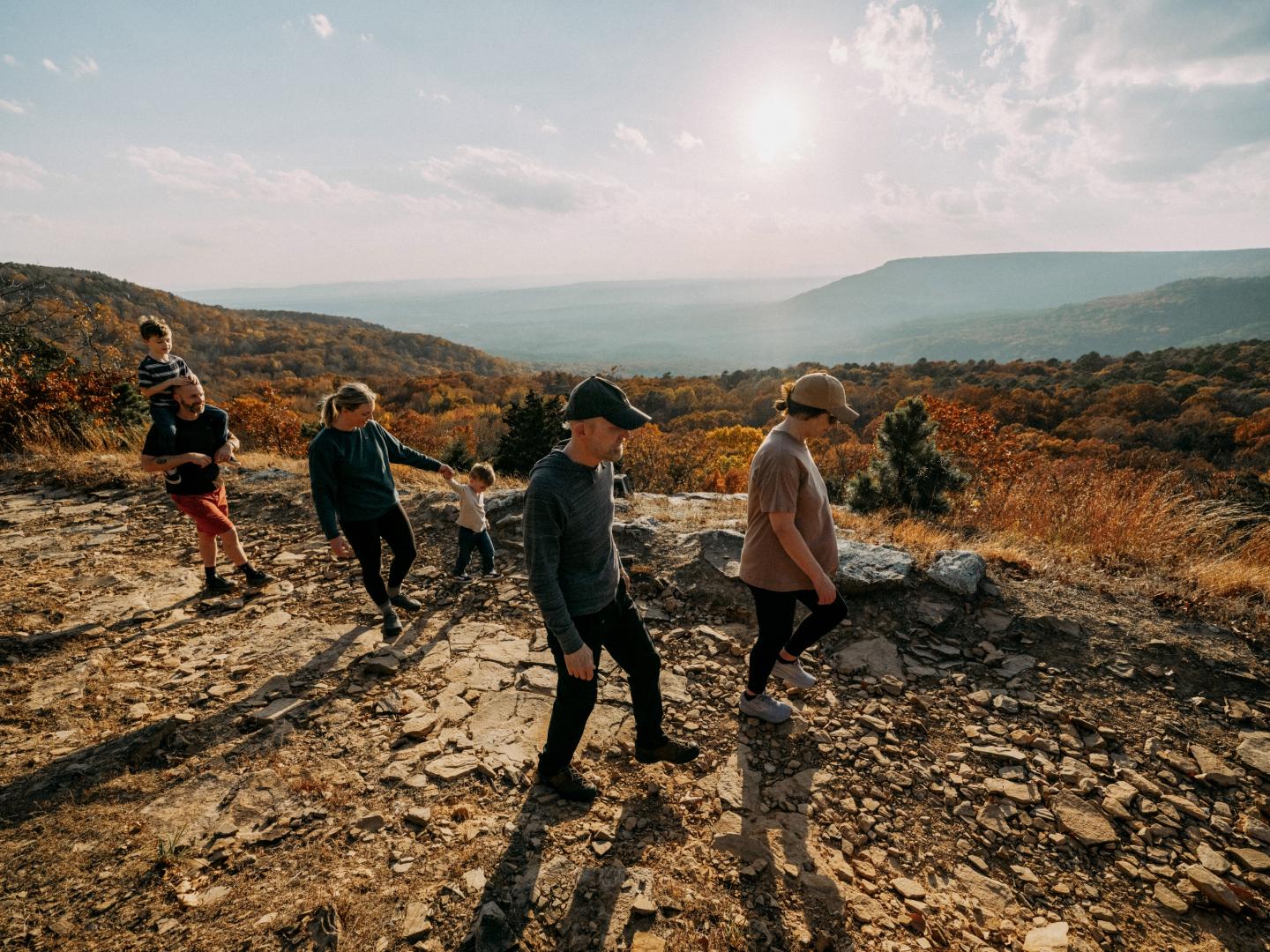 Four hikers walk along a rocky trail through sunlit hills.