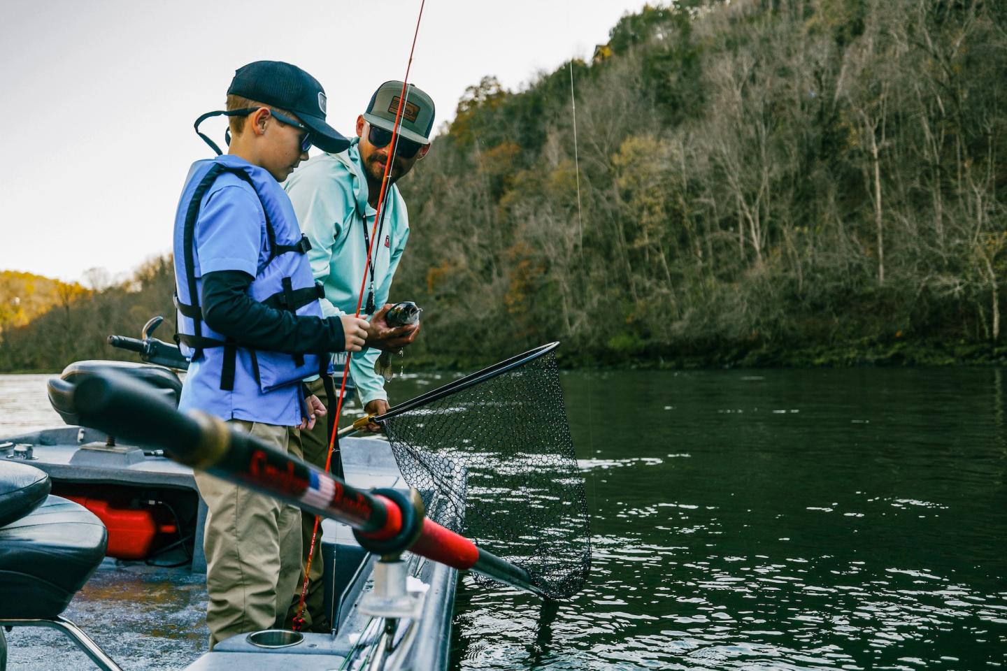 Two men fly-fishing from a boat on a river beside a forested cliff.