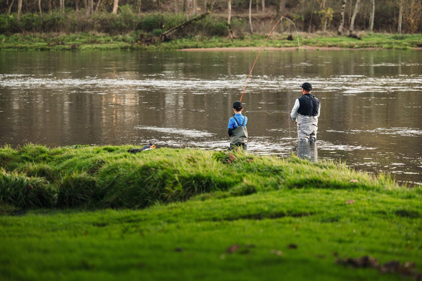 Adult and child fishing beside a calm river, viewed across green grass.