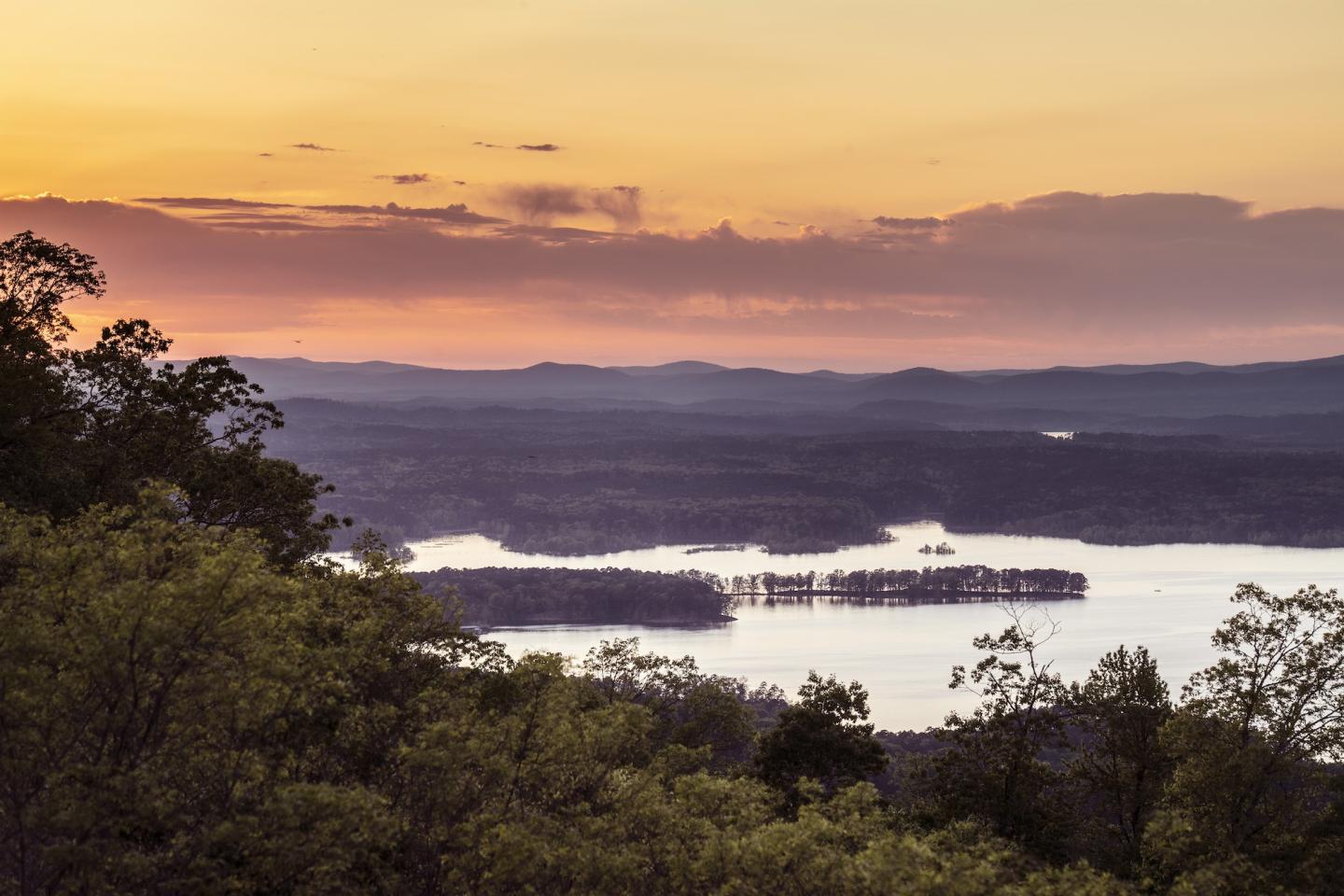 Sunset over a calm lake, framed by dark trees and distant hills.