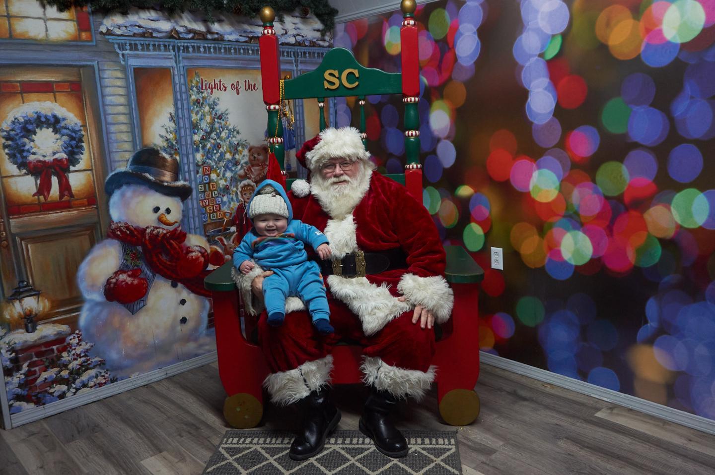 Santa and a child in blue sit beside a snowman and colorful Christmas lights.