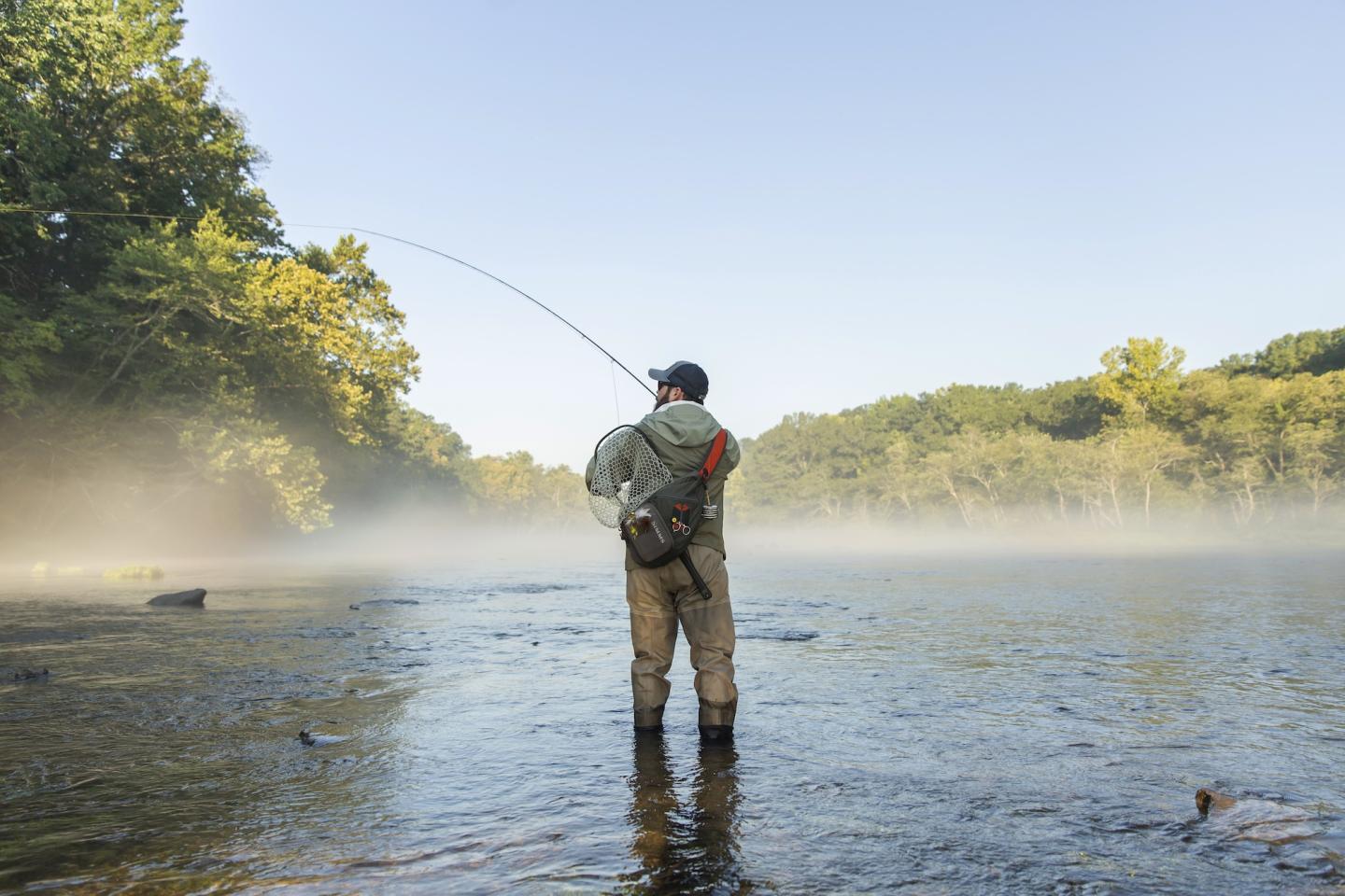 Angler standing in a misty river, casting a fishing line at sunrise.