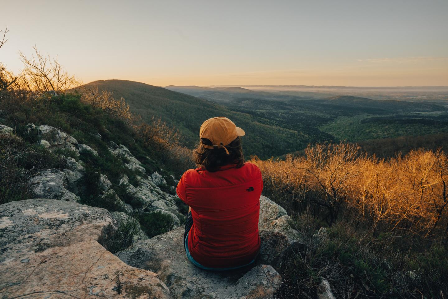 Person in a red jacket and tan hat overlooking a sunlit valley.