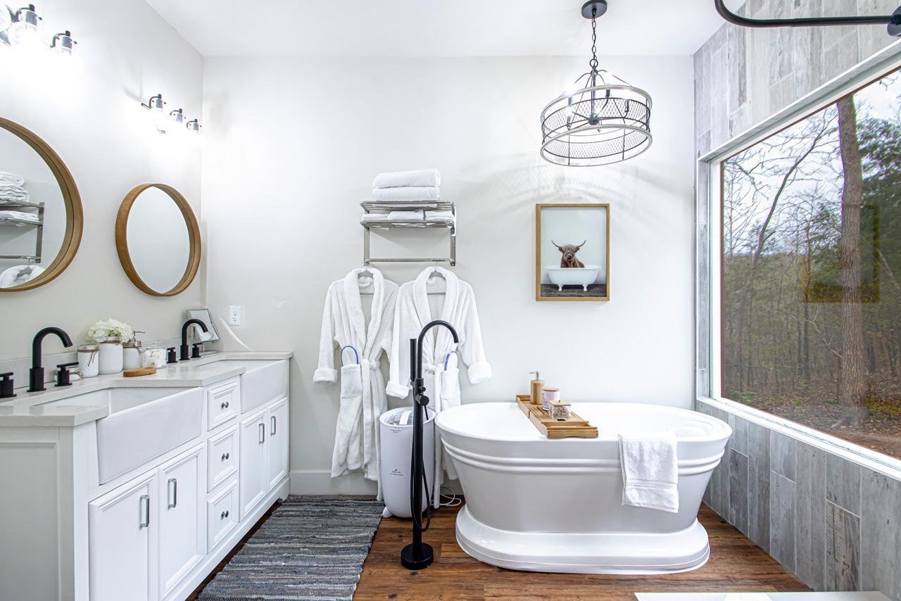 Bright white bathroom with freestanding tub, twin sinks, and large window.